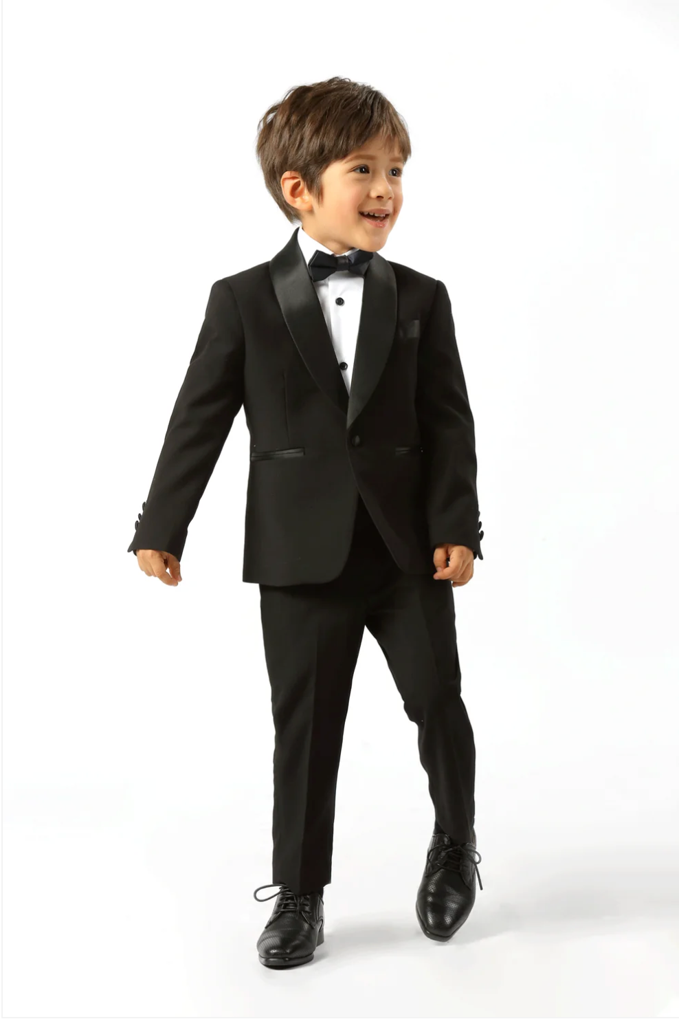 A young boy modeling a black kids' tuxedo with satin lapels, a white dress shirt, a black bow tie, and black dress shoes, photographed against a seamless white studio background.