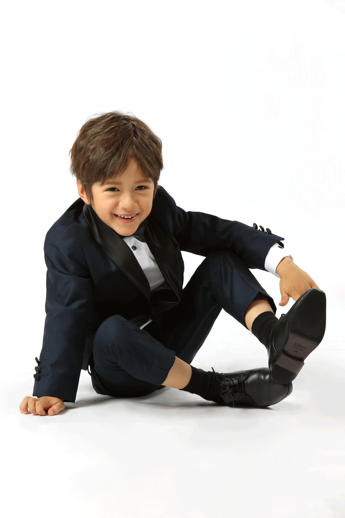 A young boy wearing a dark navy blue kids' tuxedo with black satin lapels, a white dress shirt, and black dress socks, sitting on a white studio floor.