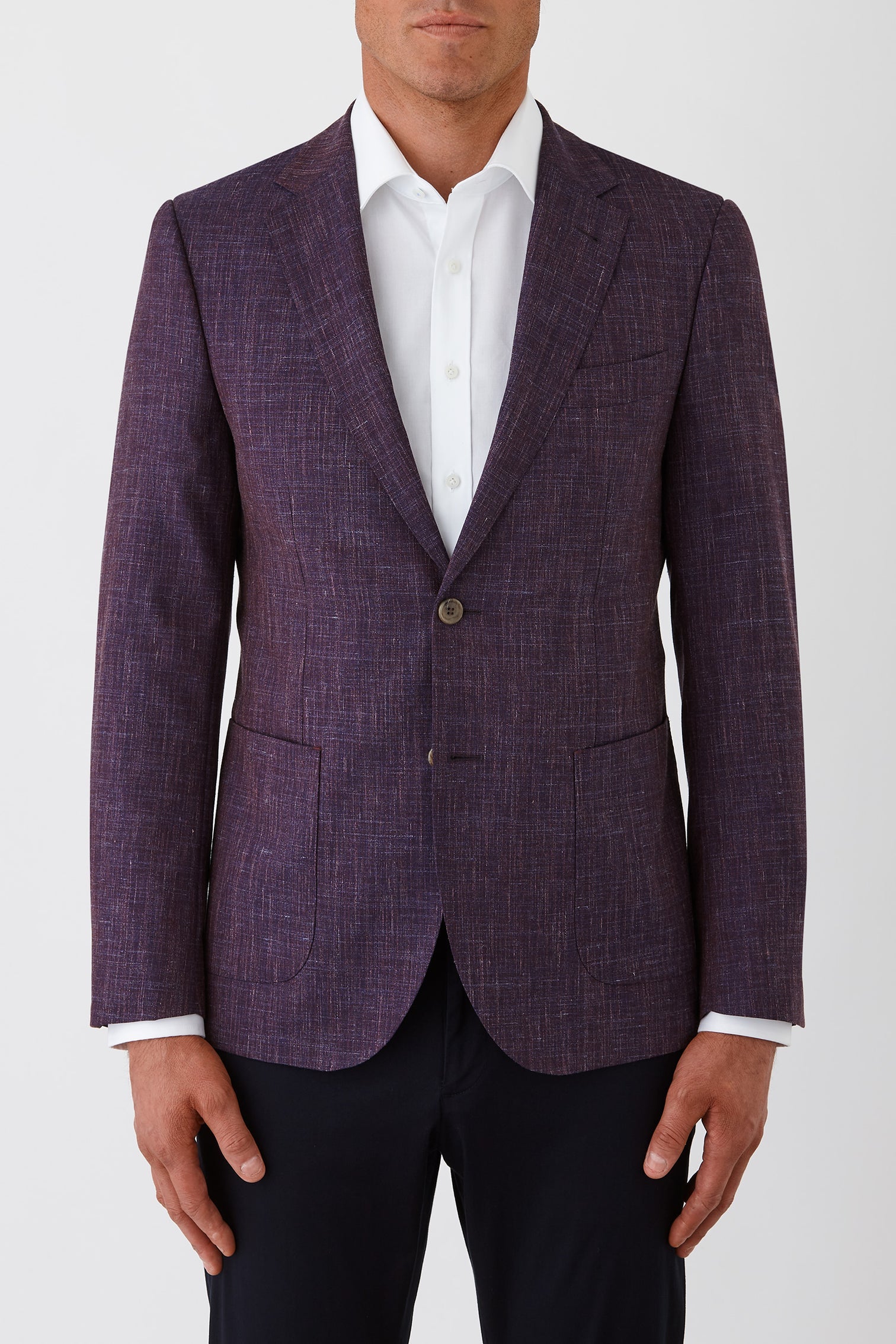 A man wearing a textured purple blazer over a white collared shirt and dark trousers, shown from the lower face to the upper thighs against a light gray studio background.