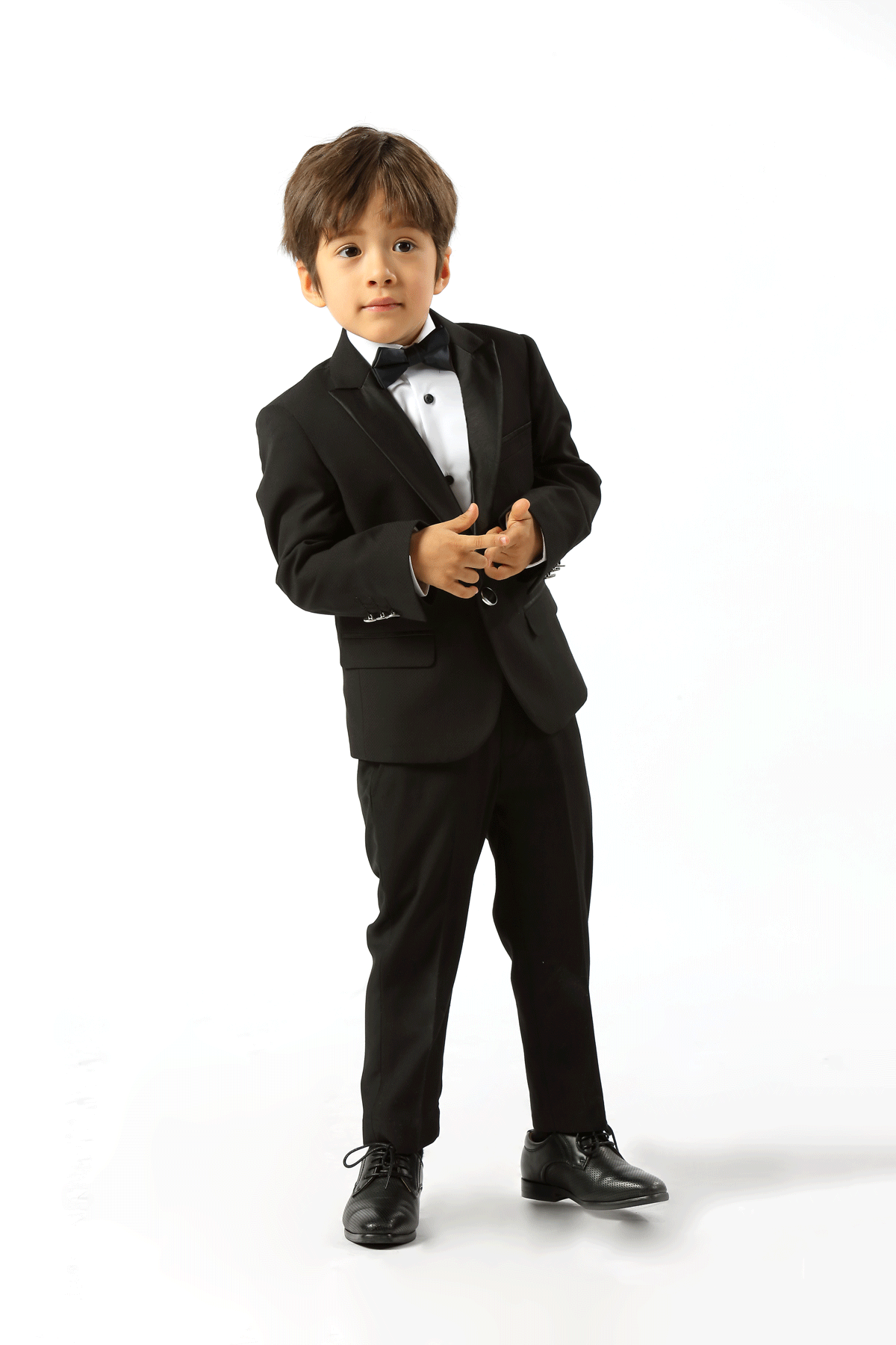 A young boy modeling a black formal tuxedo suit with a white dress shirt, black bow tie, and black dress shoes, standing against a solid white studio background.