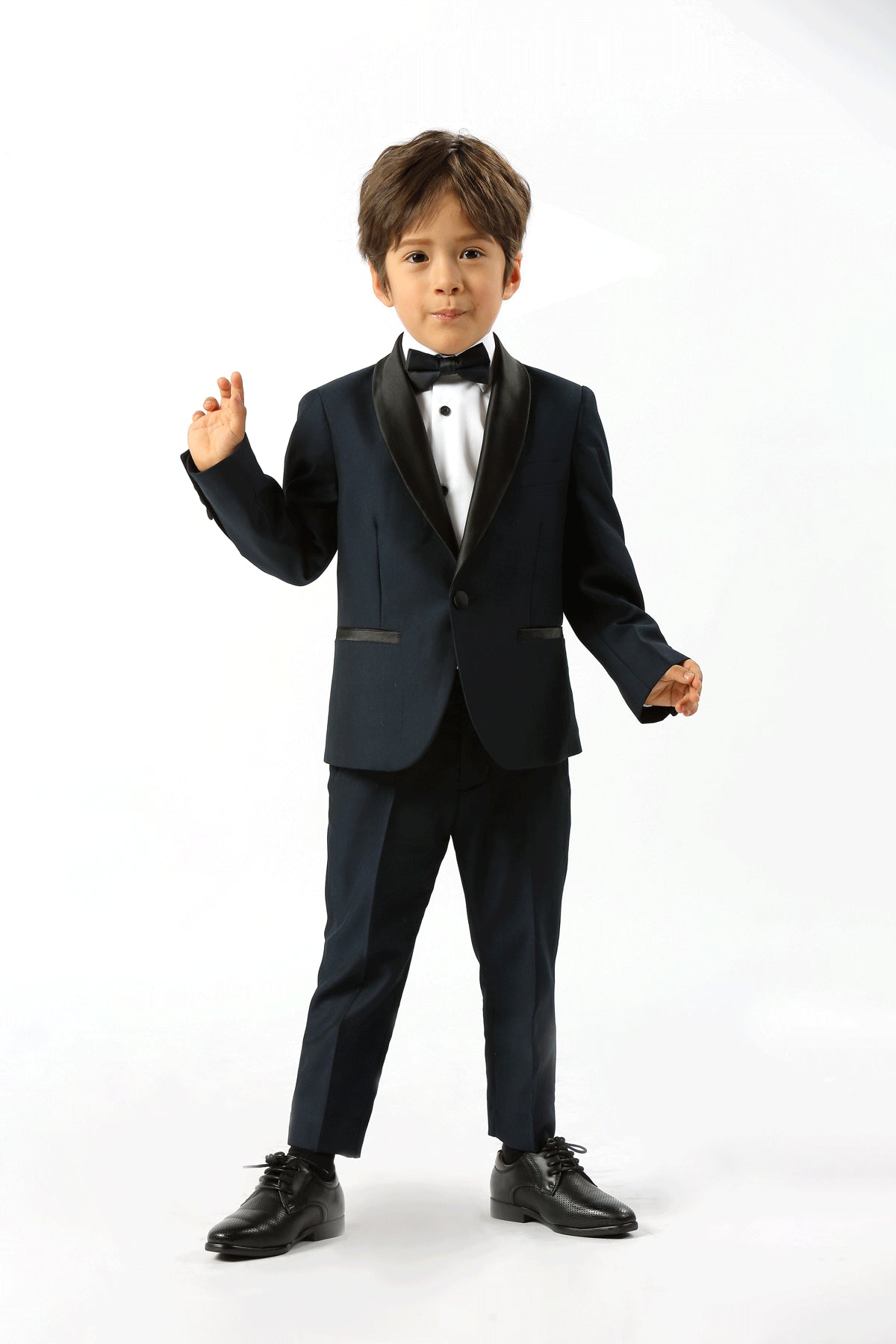 A young boy modeling a dark navy blue kids' tuxedo with black satin lapels, a white dress shirt, and a black bow tie, standing against a plain white studio background.
